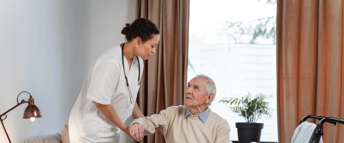 Carer in a white uniform helping an older man stand up from his bed in an aged care room