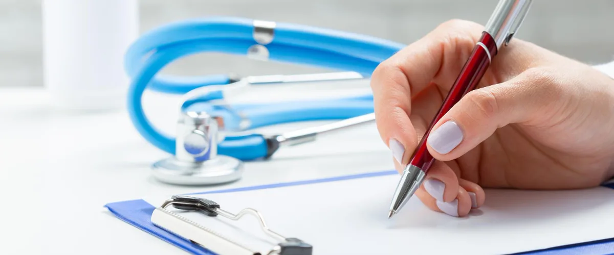Hand writing on a clipboard beside a blue stethoscope on a clinic desk