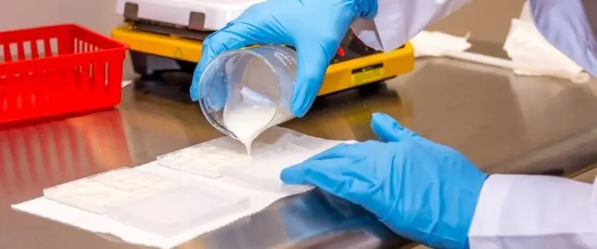 Gloved hands pouring a liquid compounded medicine into moulds on a stainless steel bench
