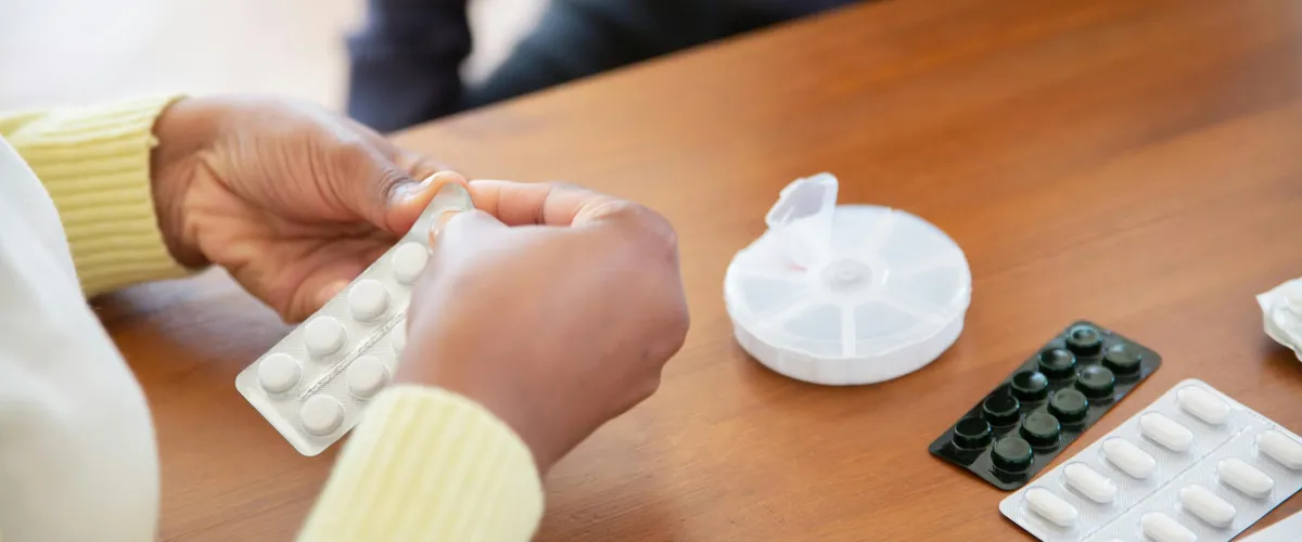 Hands pressing a tablet from a blister pack beside a pill organiser on a table